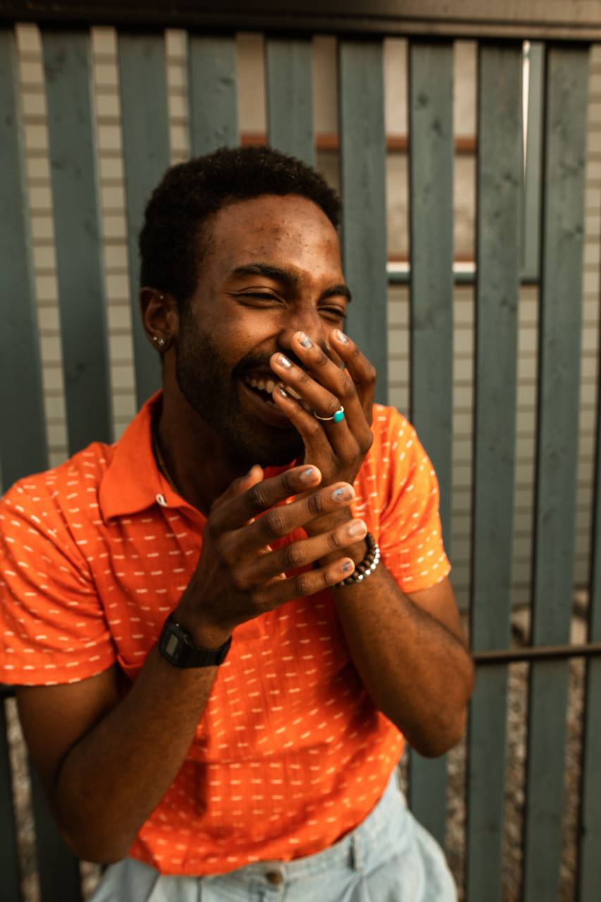 Portrait, Young Man, Smiling, Outdoors, Orange T-shirt, Afro-American, Joke, Happiness, Joyful, Laughter, Cheerful, Positive, Humor, Playful, Fun, Lighthearted, Outdoor Lifestyle, Urban, City, Confident, Carefree, Relaxed, Casual, Vibrant, Positive Vibes, Charismatic, Radiant, Sunlight, Cheerfulness, 