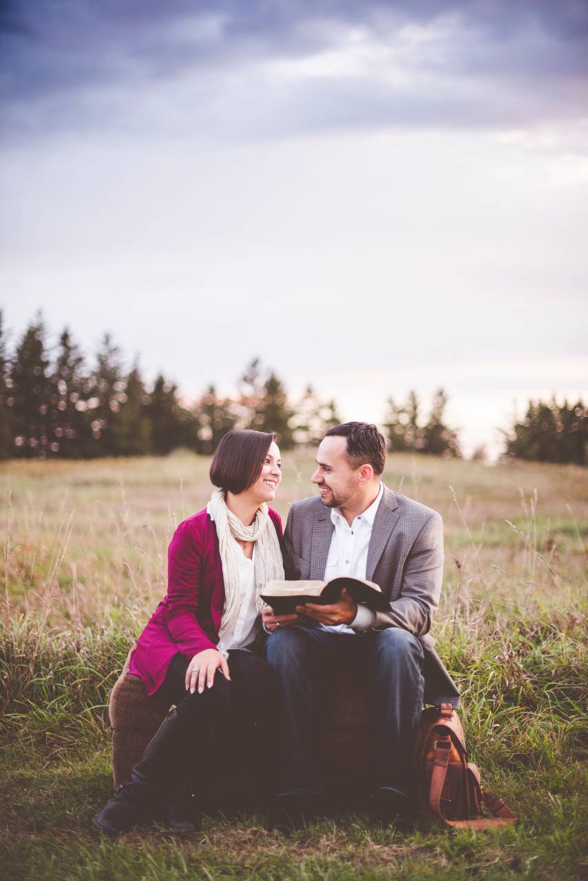 people, couple, man, woman, smile, happy, sitting, green, grass, book, bible, reading, outdoor, trees, plants, sky, clouds
, 