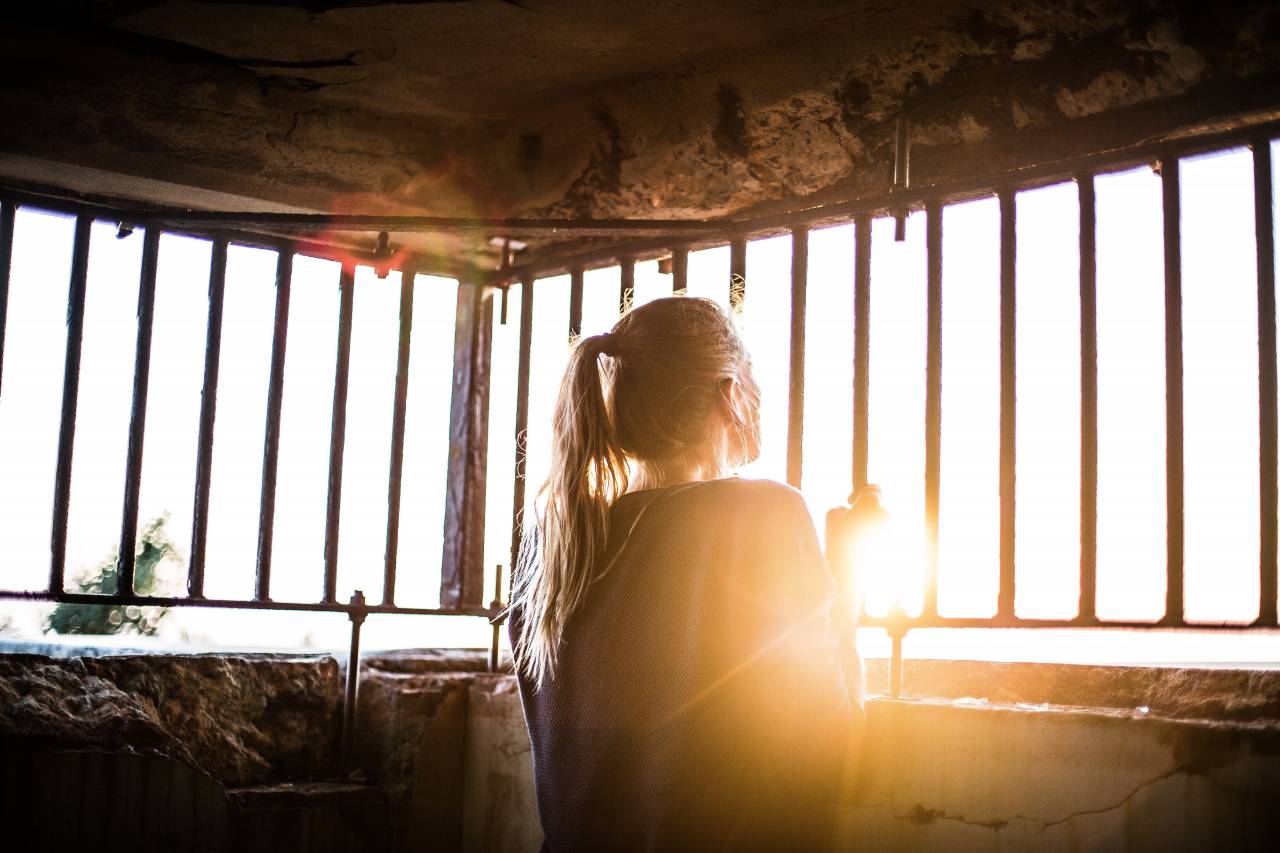 woman, girl, lady, people, back, contemplate, stand, grasp, steel, bars, concrete, room, confined, sun, light, leaks, flare
, 